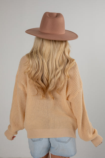 Person wearing a beige sweater and brown hat with long blonde hair, standing against a plain background