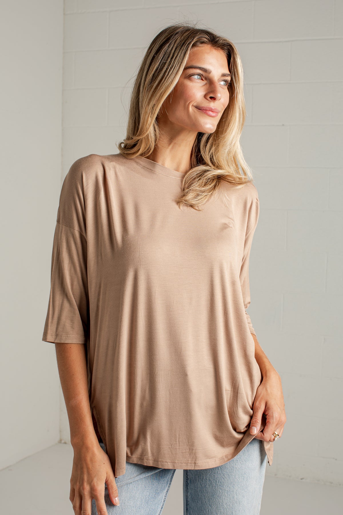 A woman wearing a loose, short-sleeve beige t-shirt and light blue jeans, standing against a white brick wall.