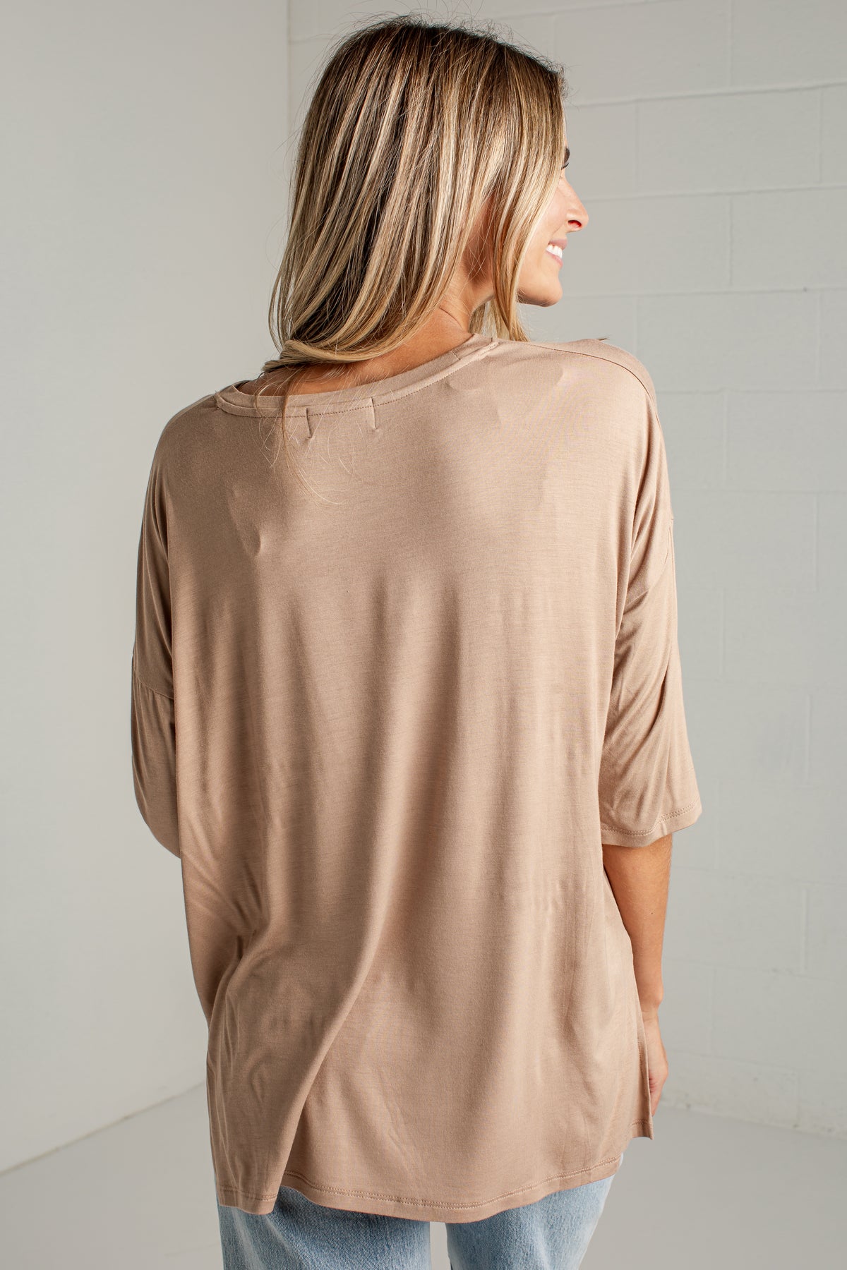A woman shown from the back wearing a loose, short-sleeve beige top and light blue jeans, standing against a white brick wall.