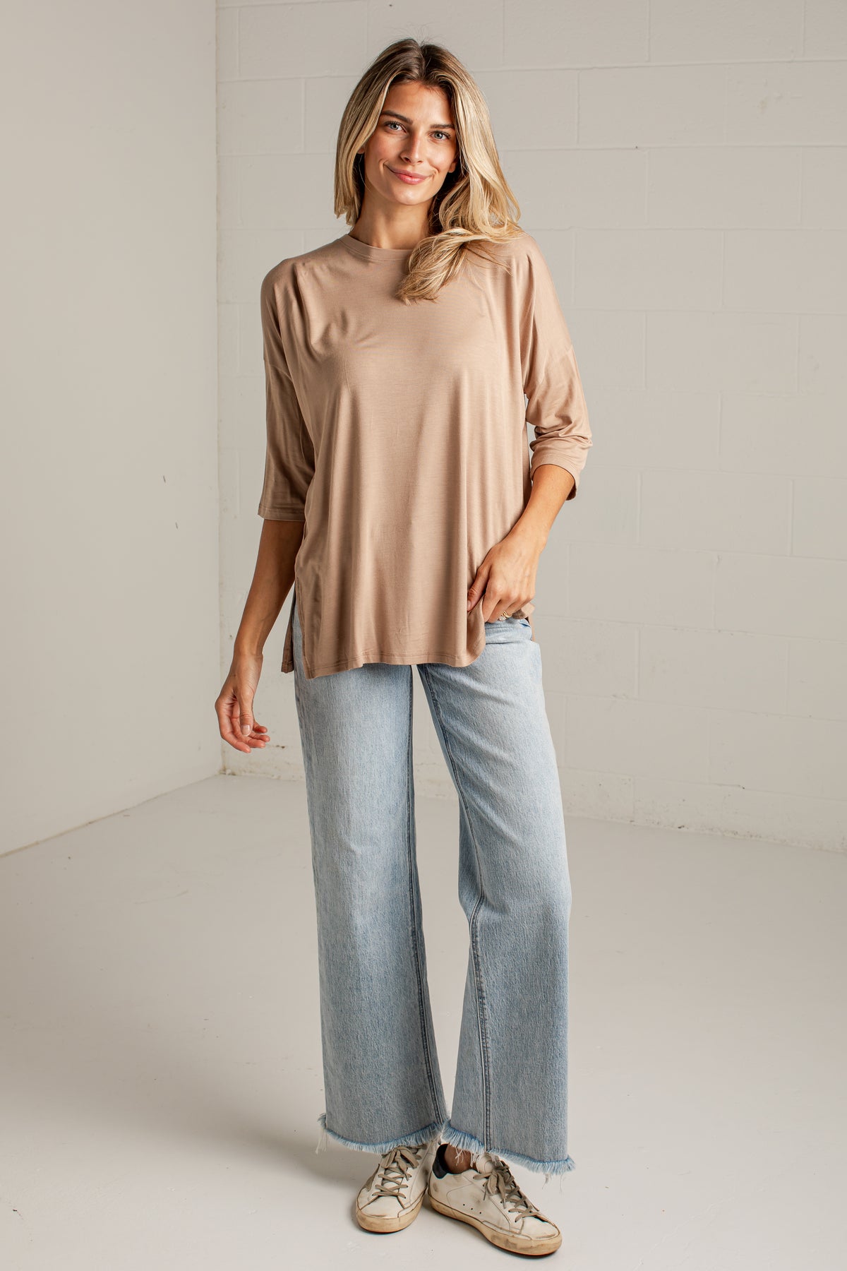 A woman modeling a beige long-sleeve top with rolled cuffs, paired with light-wash wide-leg jeans and beige sneakers, standing against a white brick wall.