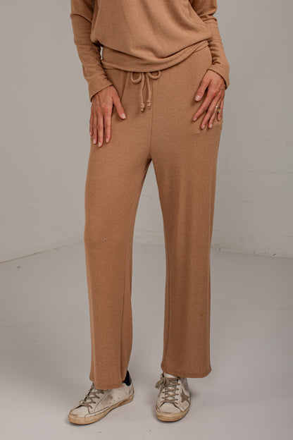 A person wearing ribbed knit wide-leg pants in a warm camel brown, paired with a matching long-sleeve top and distressed white sneakers, standing against a light gray studio background.