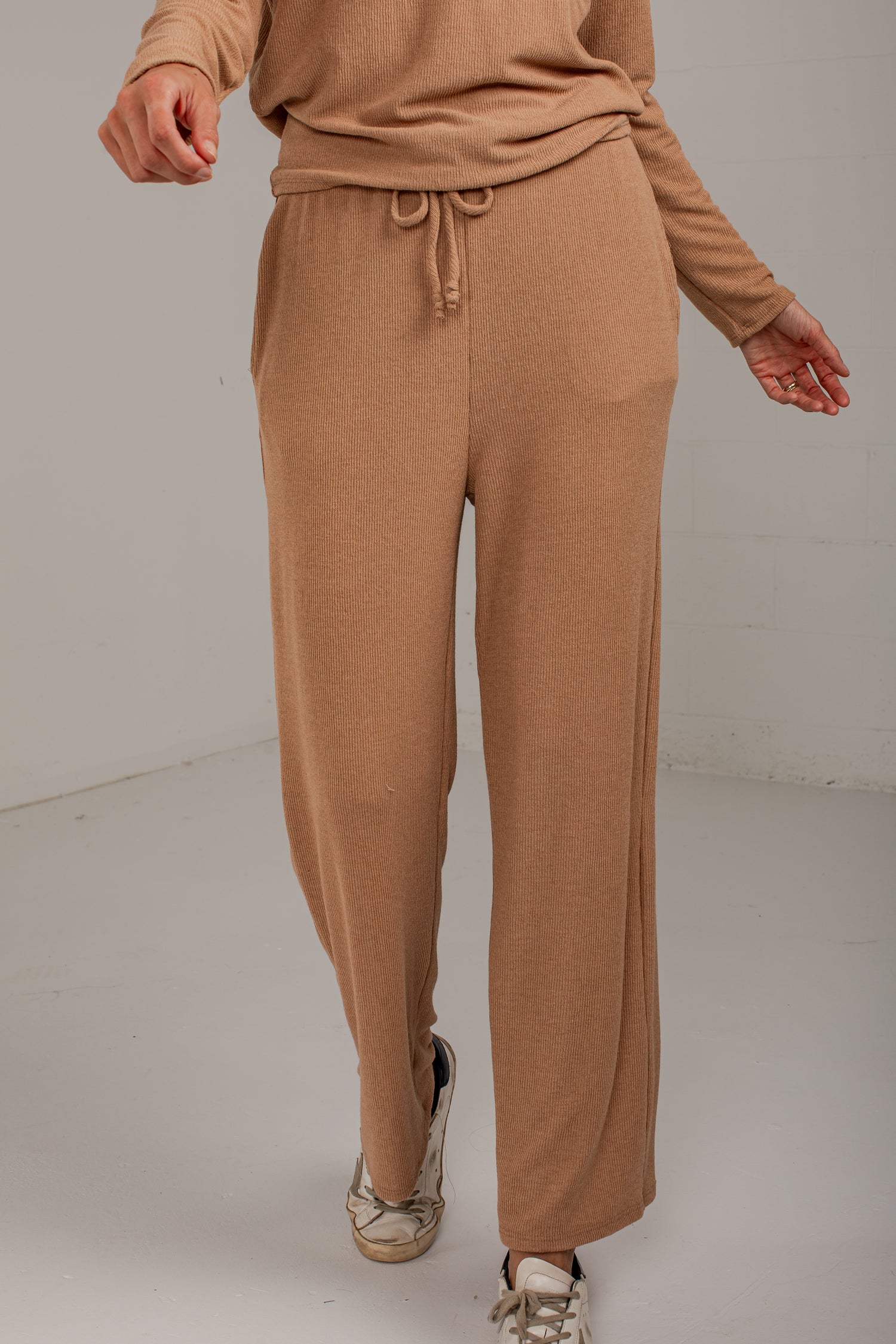 A person wearing a matching tan knit lounge set, featuring wide-leg drawstring pants and a long-sleeve top, paired with metallic sneakers, in a studio setting.