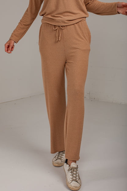 A person wearing a matching tan knit lounge set, featuring wide-leg drawstring pants and a long-sleeve top, paired with distressed white sneakers, photographed in a minimalist studio setting.