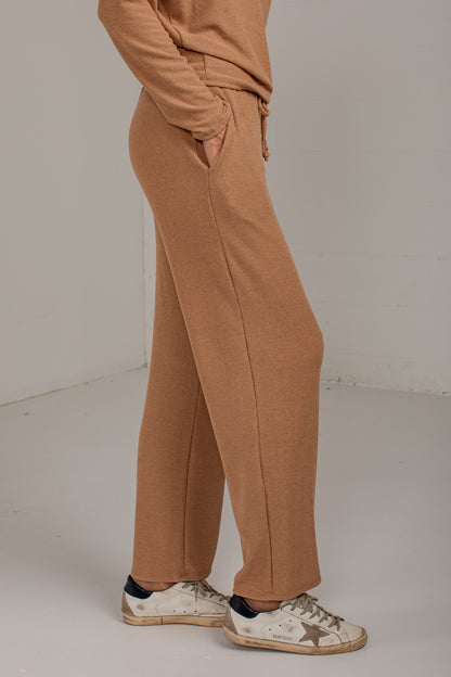A person wearing wide-leg tan sweatpants with a drawstring waist, paired with a matching tan top and white sneakers, shown from the waist down in a studio setting.