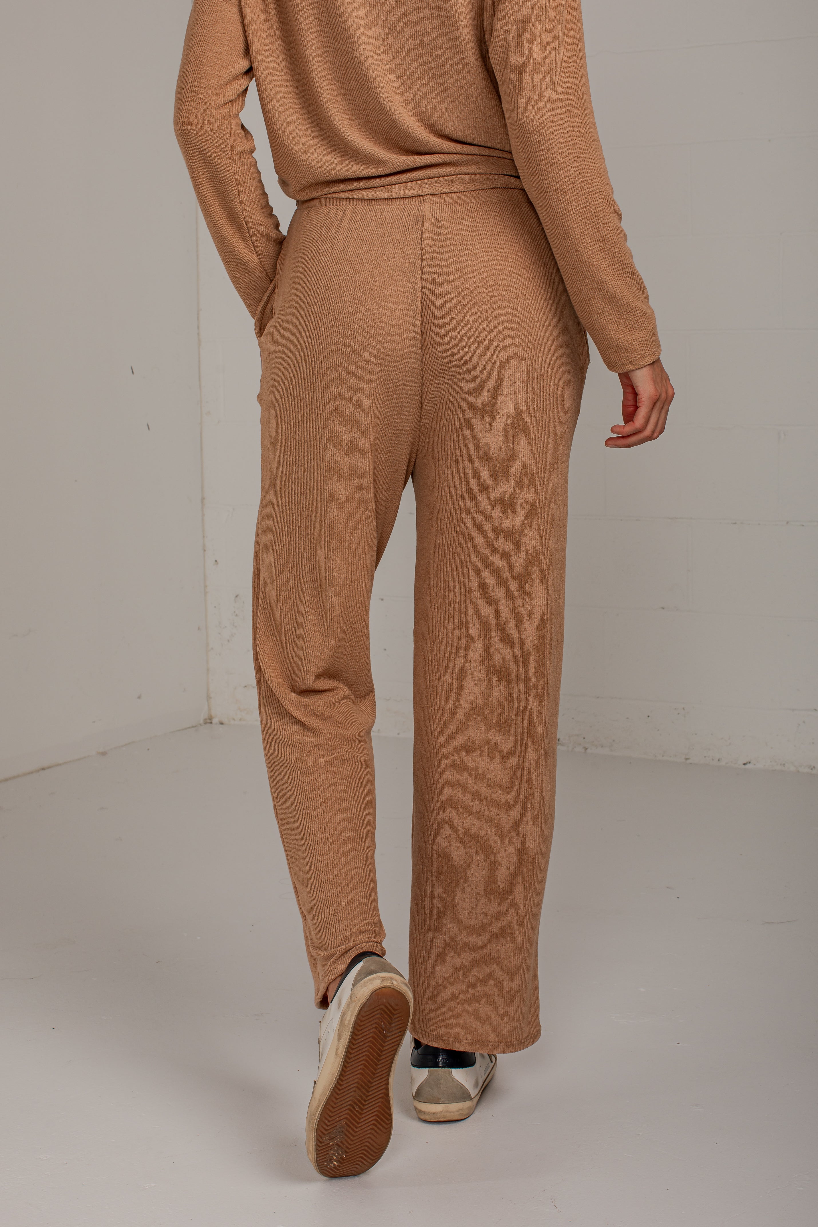 A person shown from the back wearing a matching ribbed knit two-piece set in a warm camel brown, featuring a long-sleeve top and wide-leg pants, paired with white sneakers.