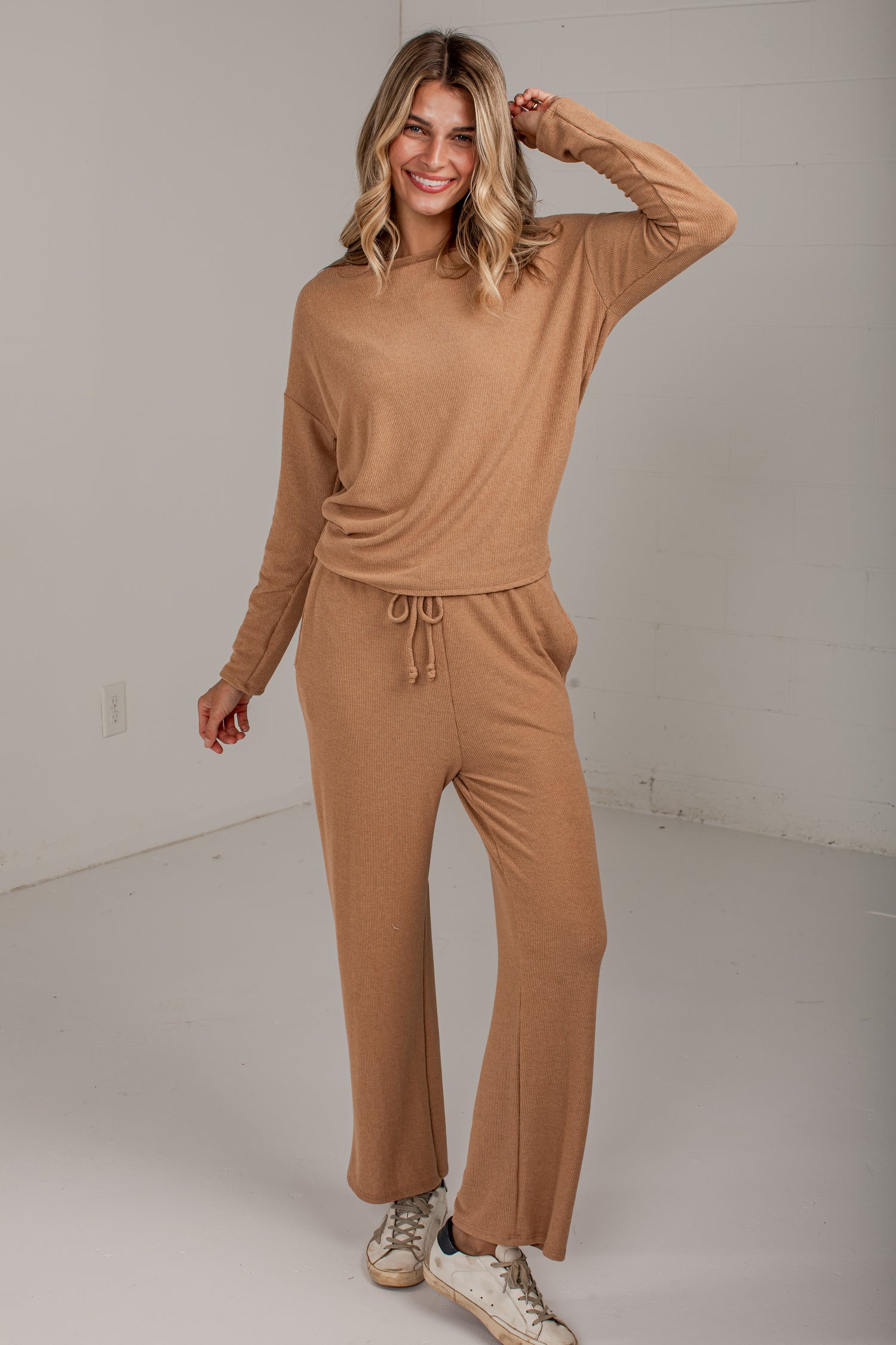 A woman modeling a matching two-piece ribbed knit loungewear set in a warm camel tan color, consisting of a long-sleeve crewneck top and wide-leg drawstring pants, paired with white sneakers in a bright studio setting.