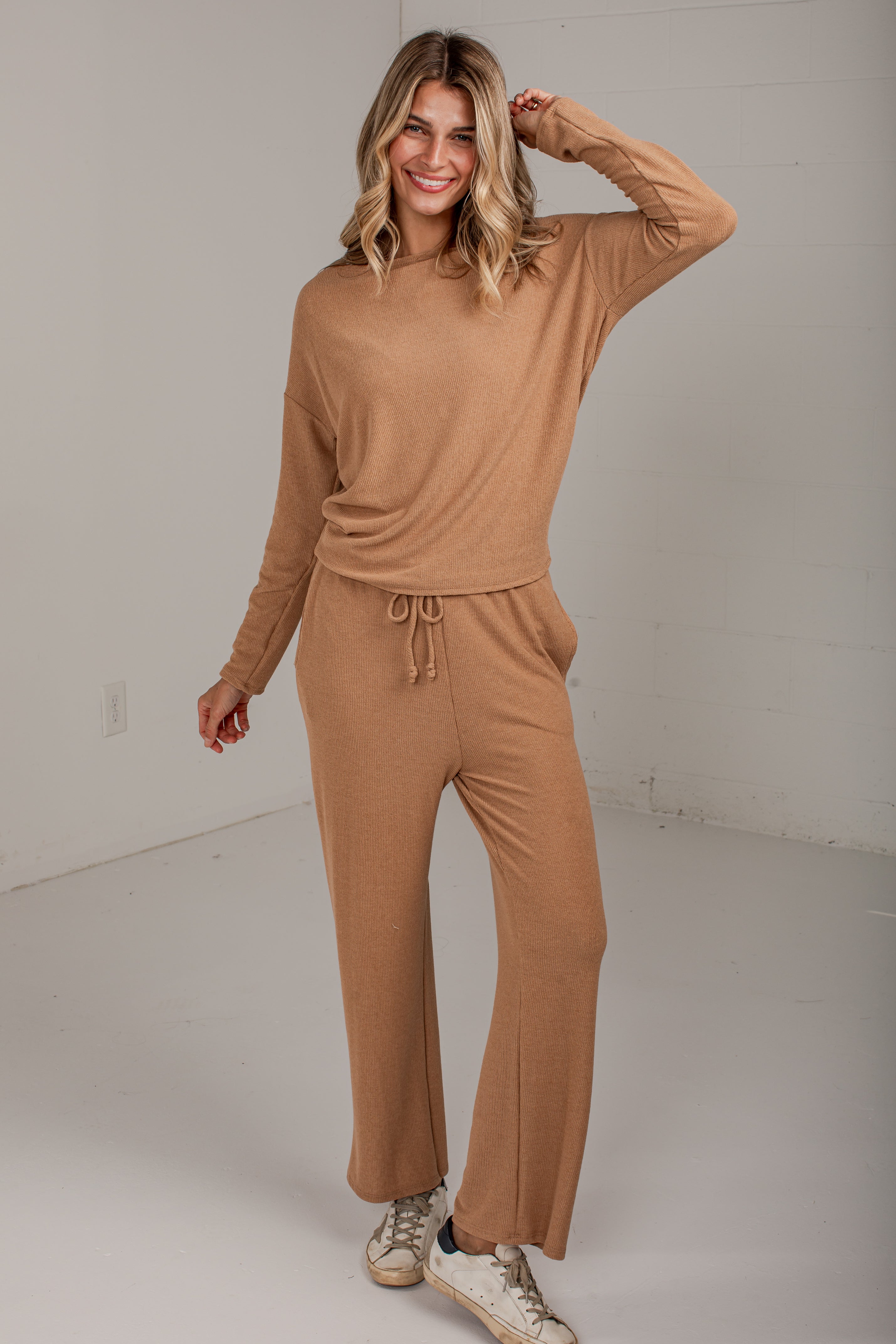 A woman modeling a matching two-piece ribbed knit loungewear set in a warm camel tan color, consisting of a long-sleeve crewneck top and wide-leg drawstring pants, paired with white sneakers in a bright studio setting.