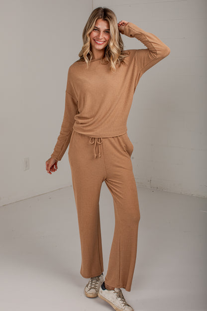 A woman modeling a matching two-piece ribbed knit loungewear set in a warm camel tan color, consisting of a long-sleeve crewneck top and wide-leg drawstring pants, paired with white sneakers in a bright studio setting.