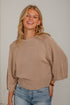 A smiling blonde woman wearing a ribbed knit beige sweater with wide three-quarter sleeves and light blue jeans, standing against a light gray wall.