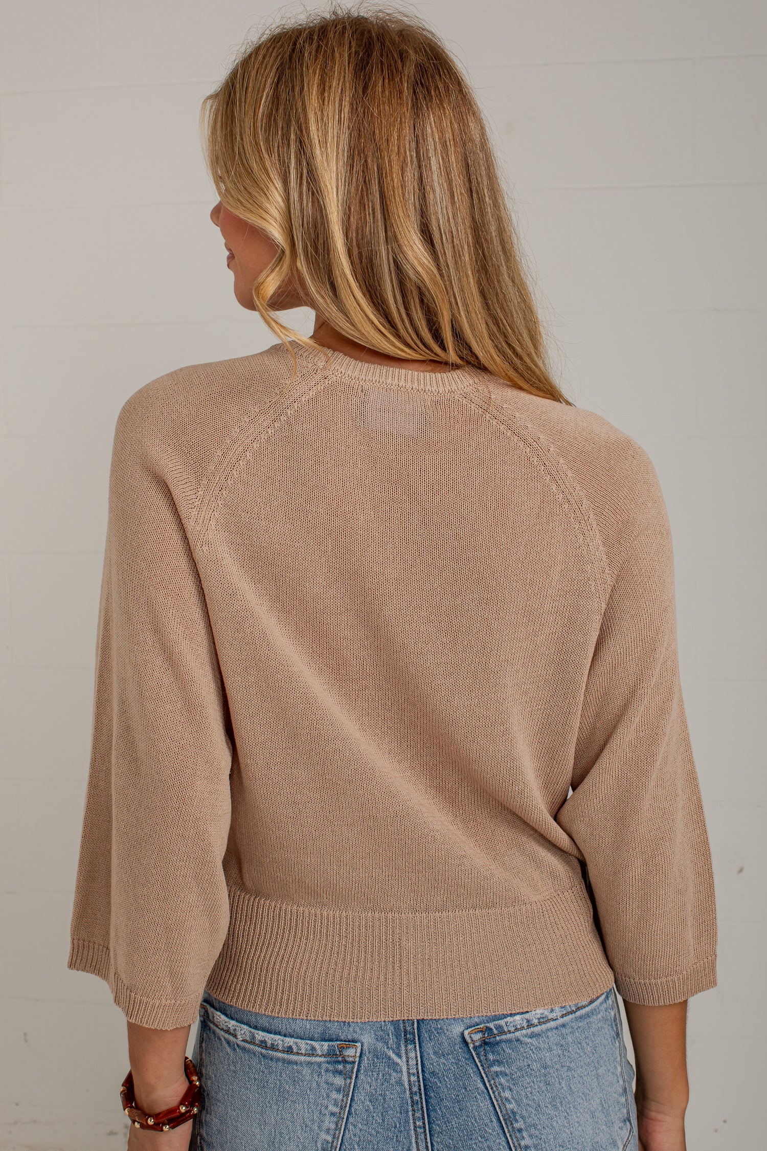 A woman viewed from behind wearing a light tan knit sweater with three-quarter sleeves and blue jeans, standing against a white paneled wall.