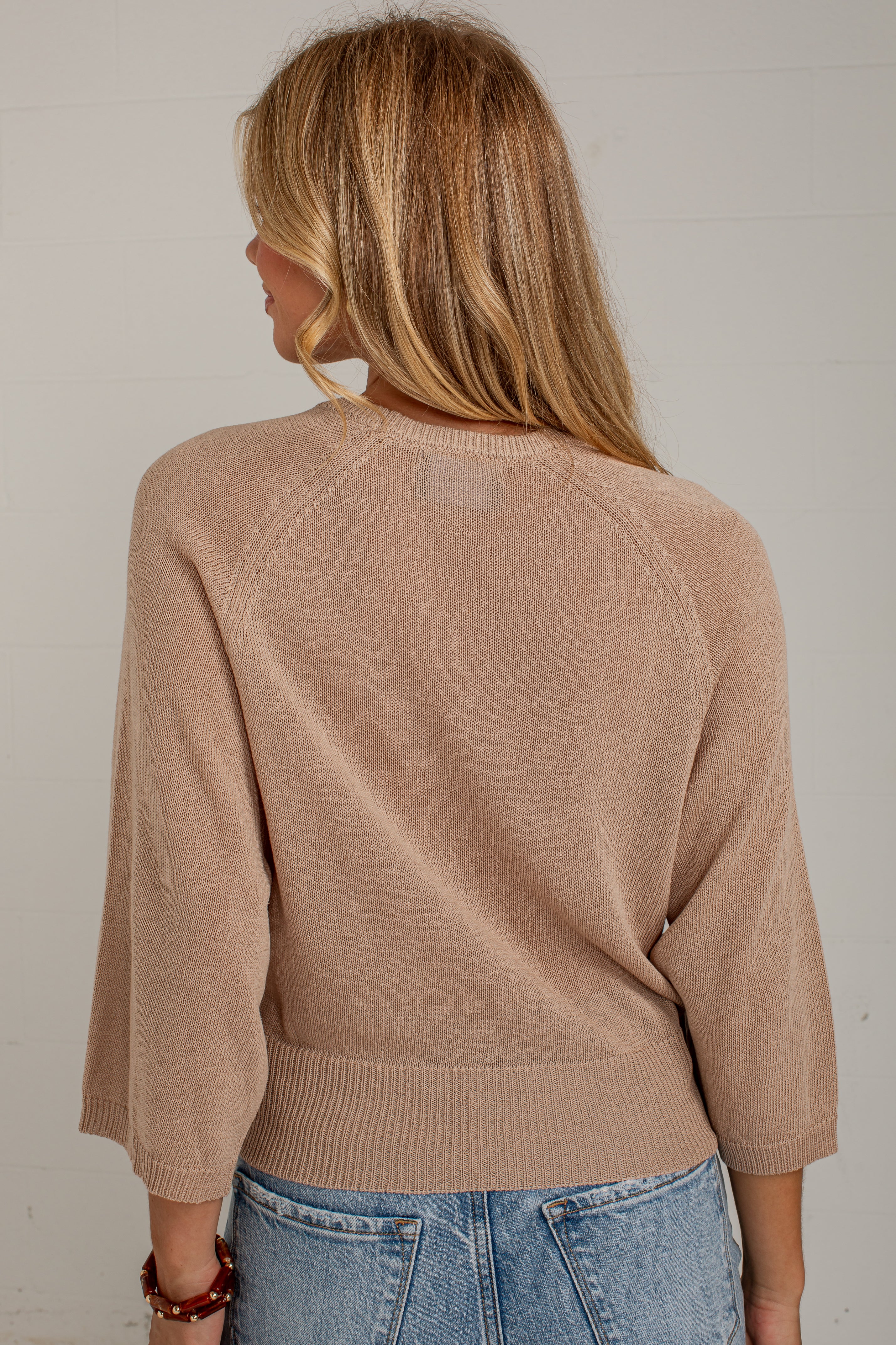 A woman viewed from behind wearing a light tan knit sweater with three-quarter sleeves and blue jeans, standing against a white paneled wall.