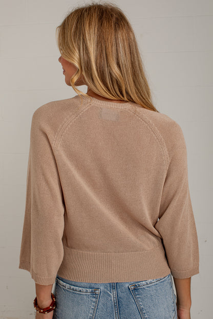 A woman viewed from behind wearing a light tan knit sweater with three-quarter sleeves and blue jeans, standing against a white paneled wall.