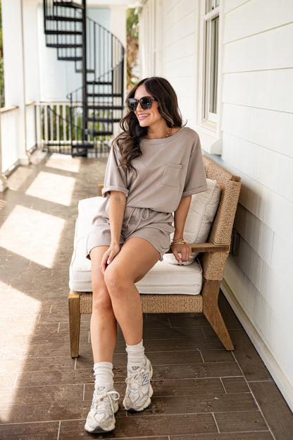 Woman sitting on a chair wearing a beige outfit with sunglasses on a sunlit porch.