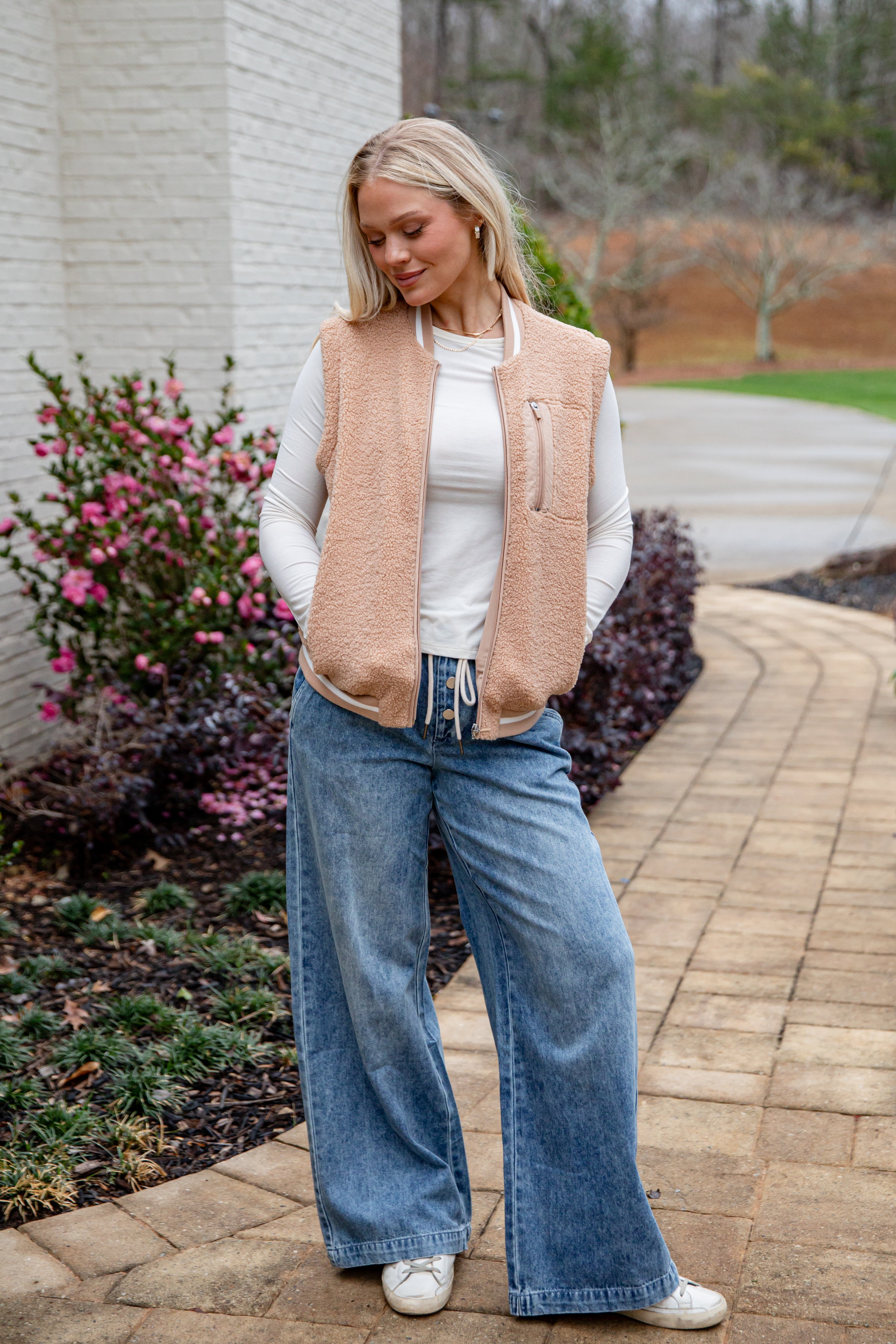 Woman wearing a beige vest, white shirt, and blue jeans standing outdoors.