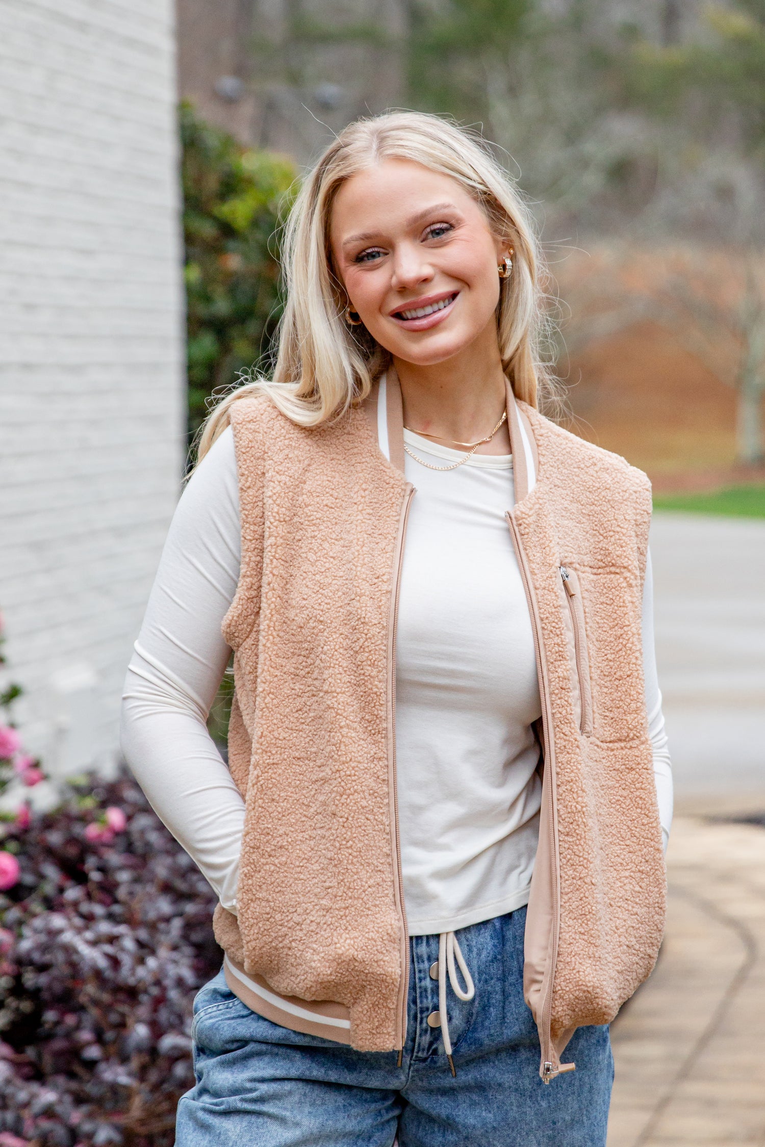 Woman wearing a beige vest over a white shirt with blue jeans, standing outdoors.