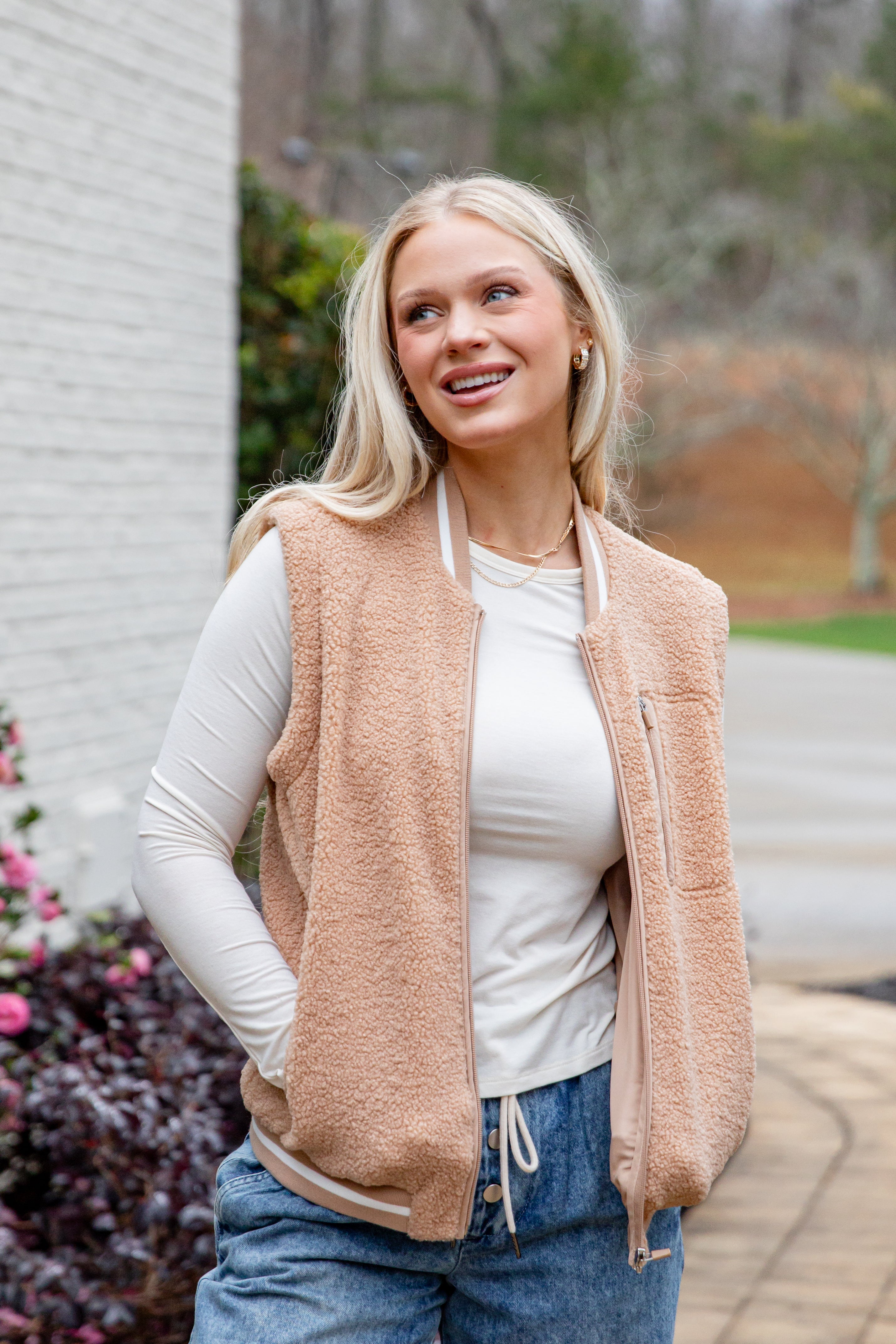 Woman wearing a beige vest over a white shirt and blue jeans, standing outdoors.