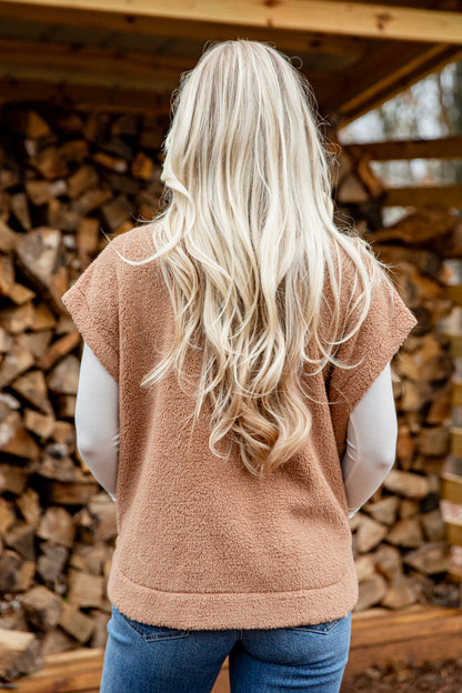 Person wearing a brown vest standing in front of stacked firewood.