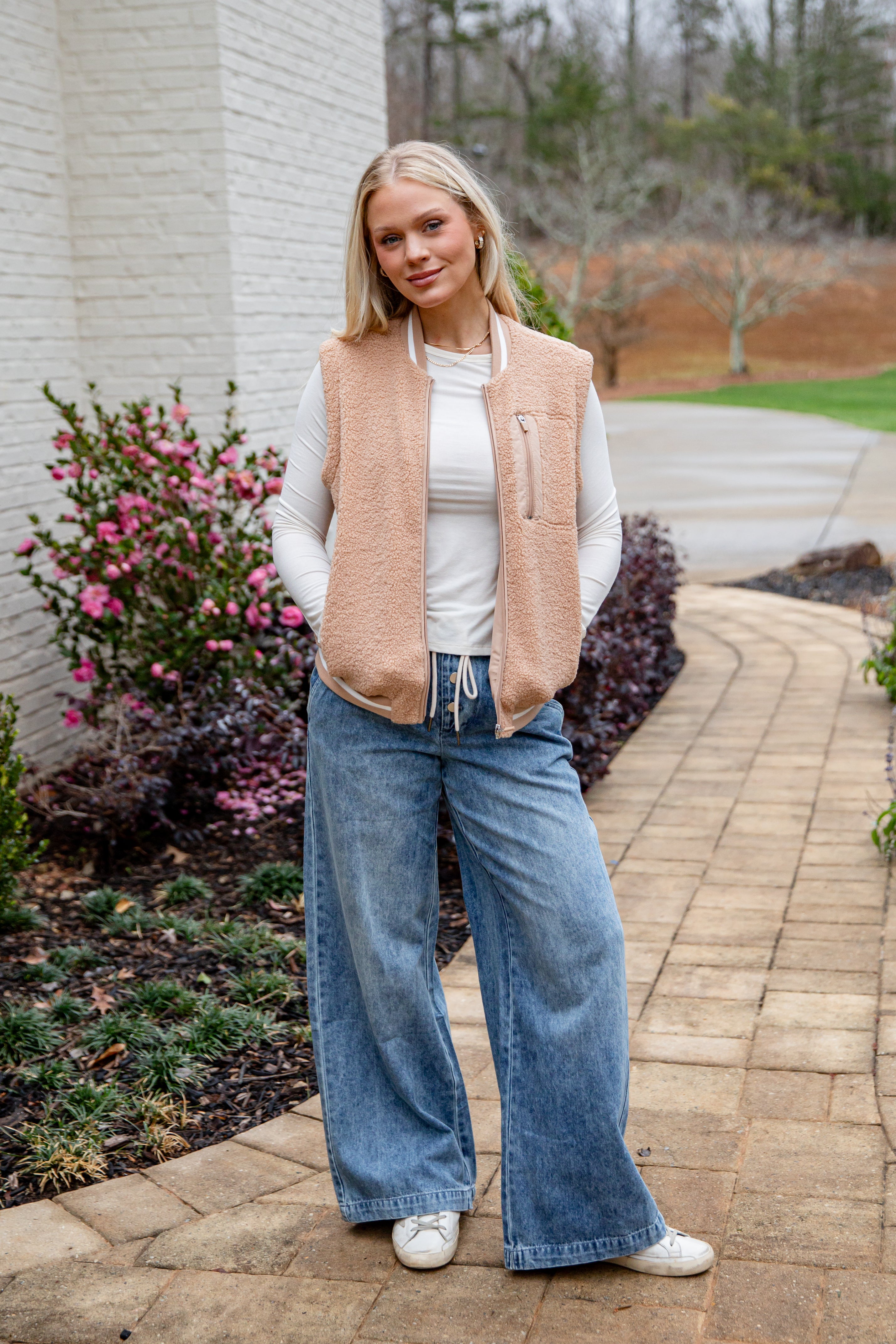 Woman wearing a beige vest, white shirt, blue jeans, and white sneakers standing on a brick path with plants and flowers in the background.