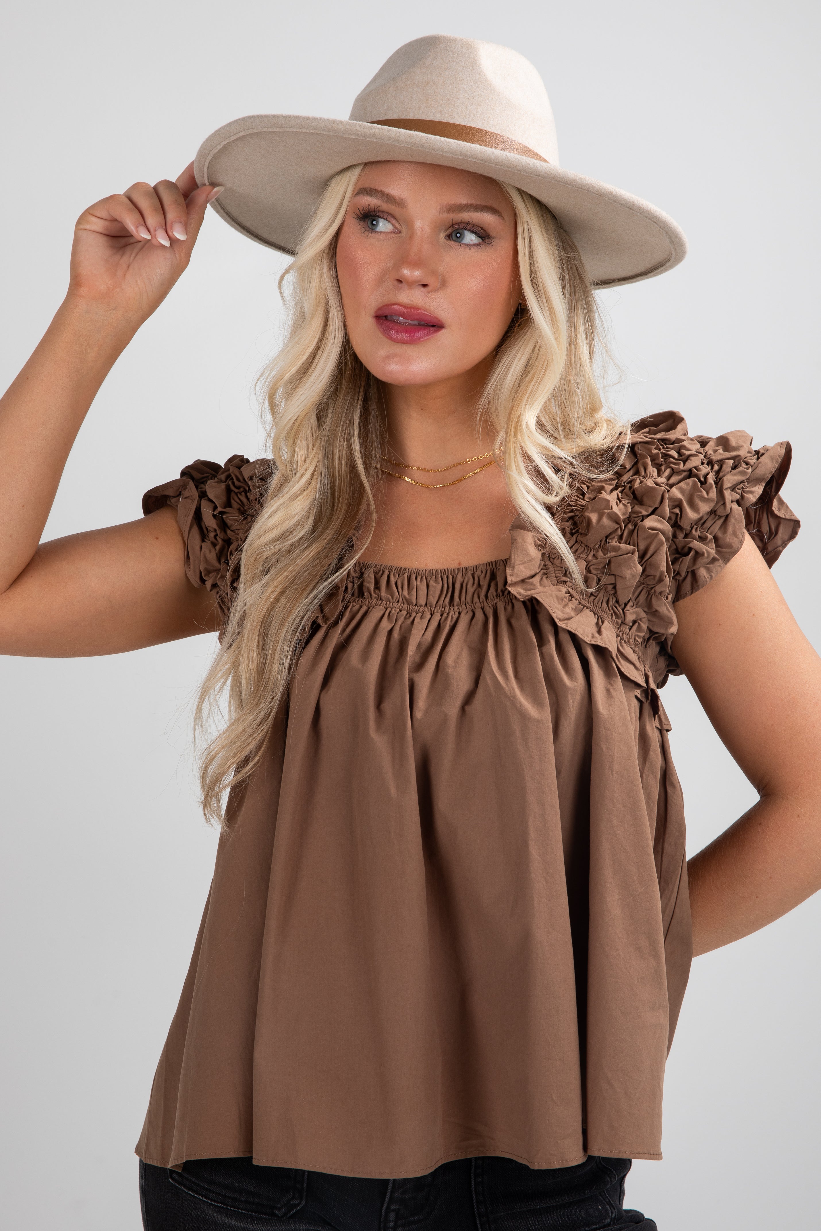 Woman wearing a brown top with ruffled sleeves and a beige hat on a white background