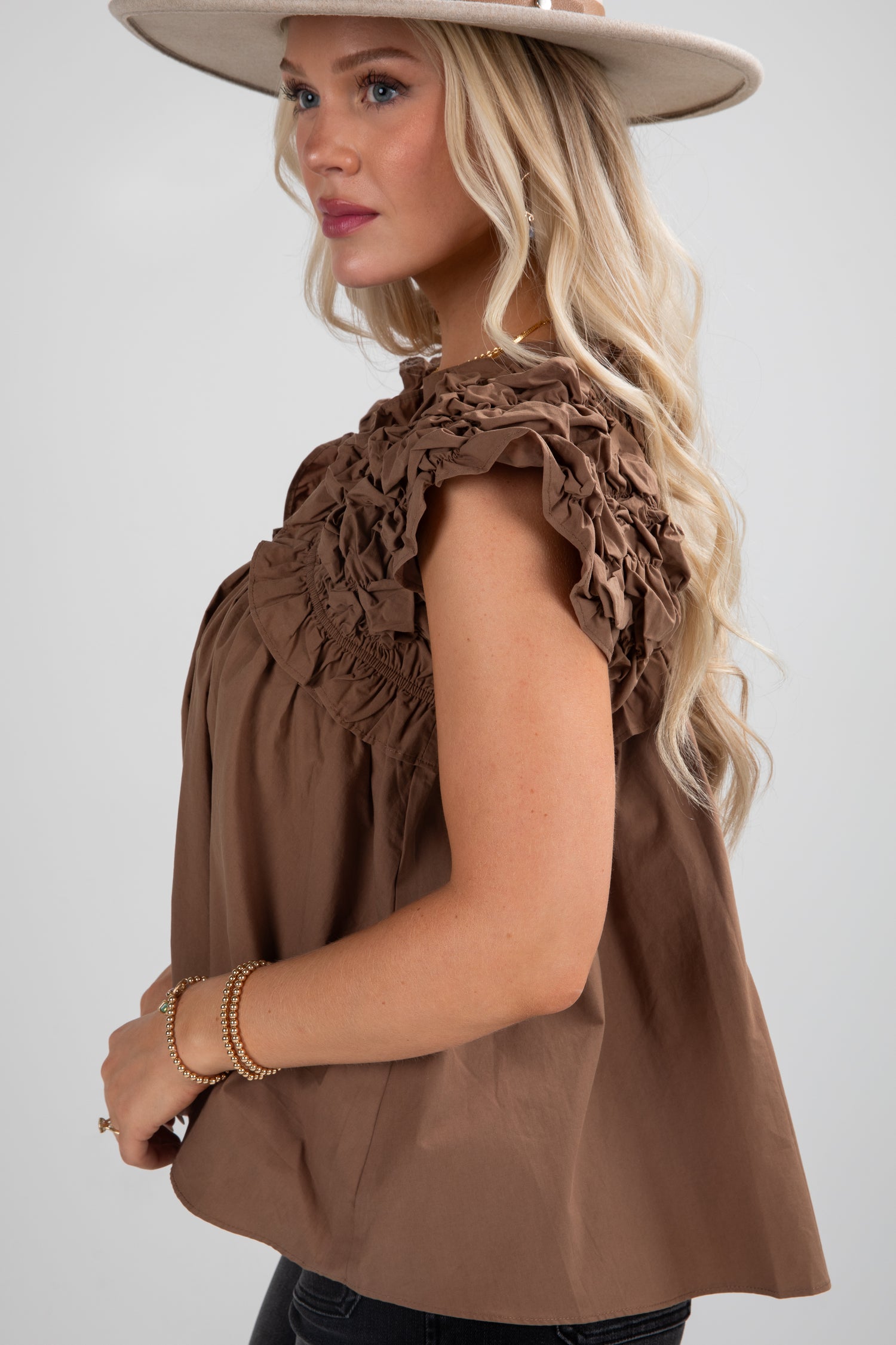 Woman wearing a brown sleeveless top with ruffled details and a beige hat on a white background