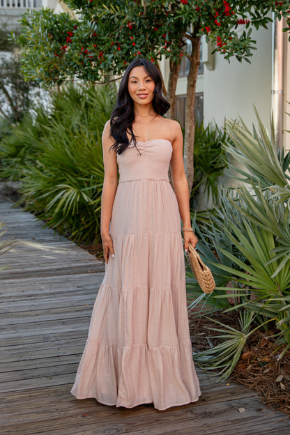 Woman in a long, light-colored dress standing on a wooden path with greenery around.