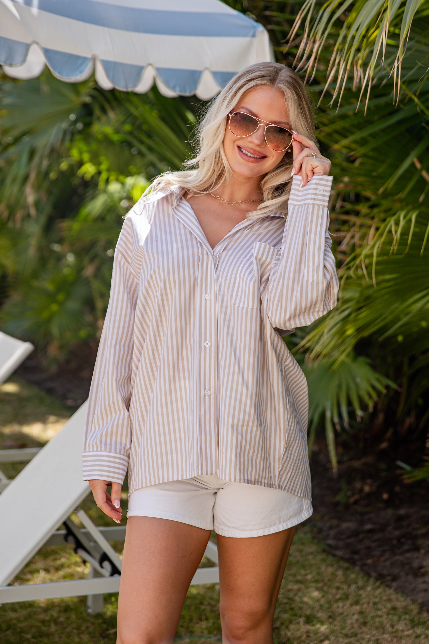 Woman in a striped shirt and shorts standing under a blue and white umbrella with greenery in the background