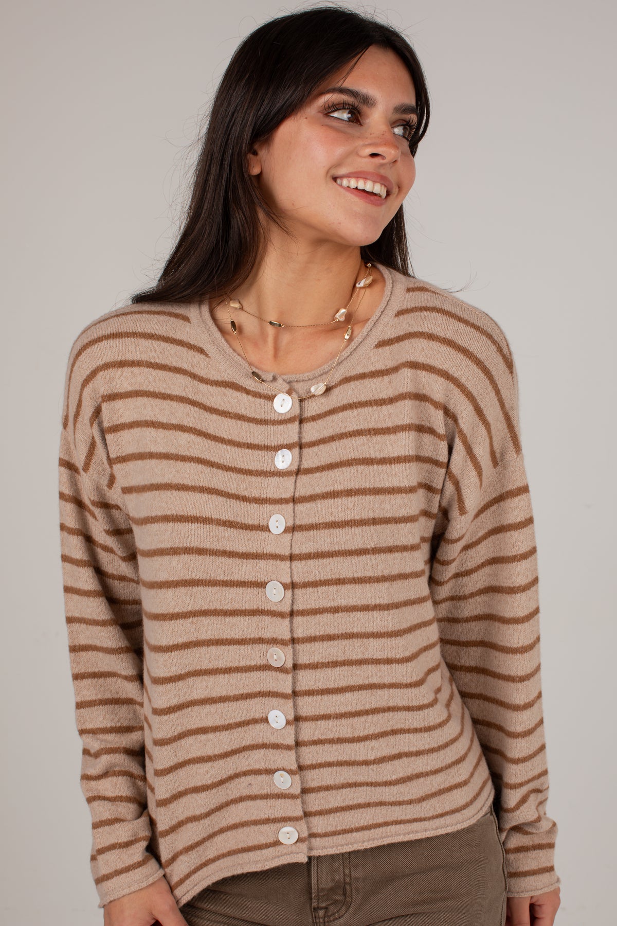 A woman wearing a beige and brown horizontally striped button-up cardigan sweater, paired with brown pants, photographed against a neutral light gray studio background.