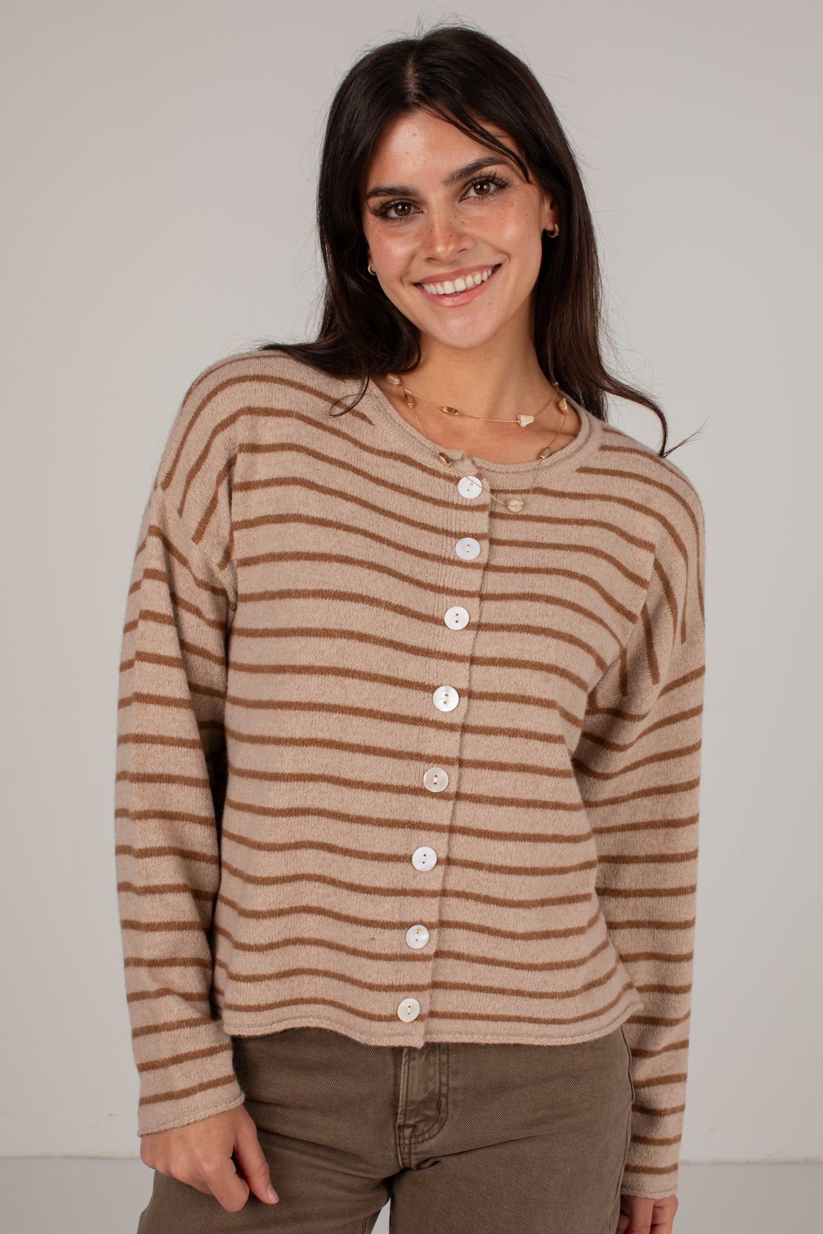 A woman wearing a cropped, long-sleeve knit cardigan with horizontal brown and beige stripes and a button-up front, paired with brown pants, against a plain light gray studio background.