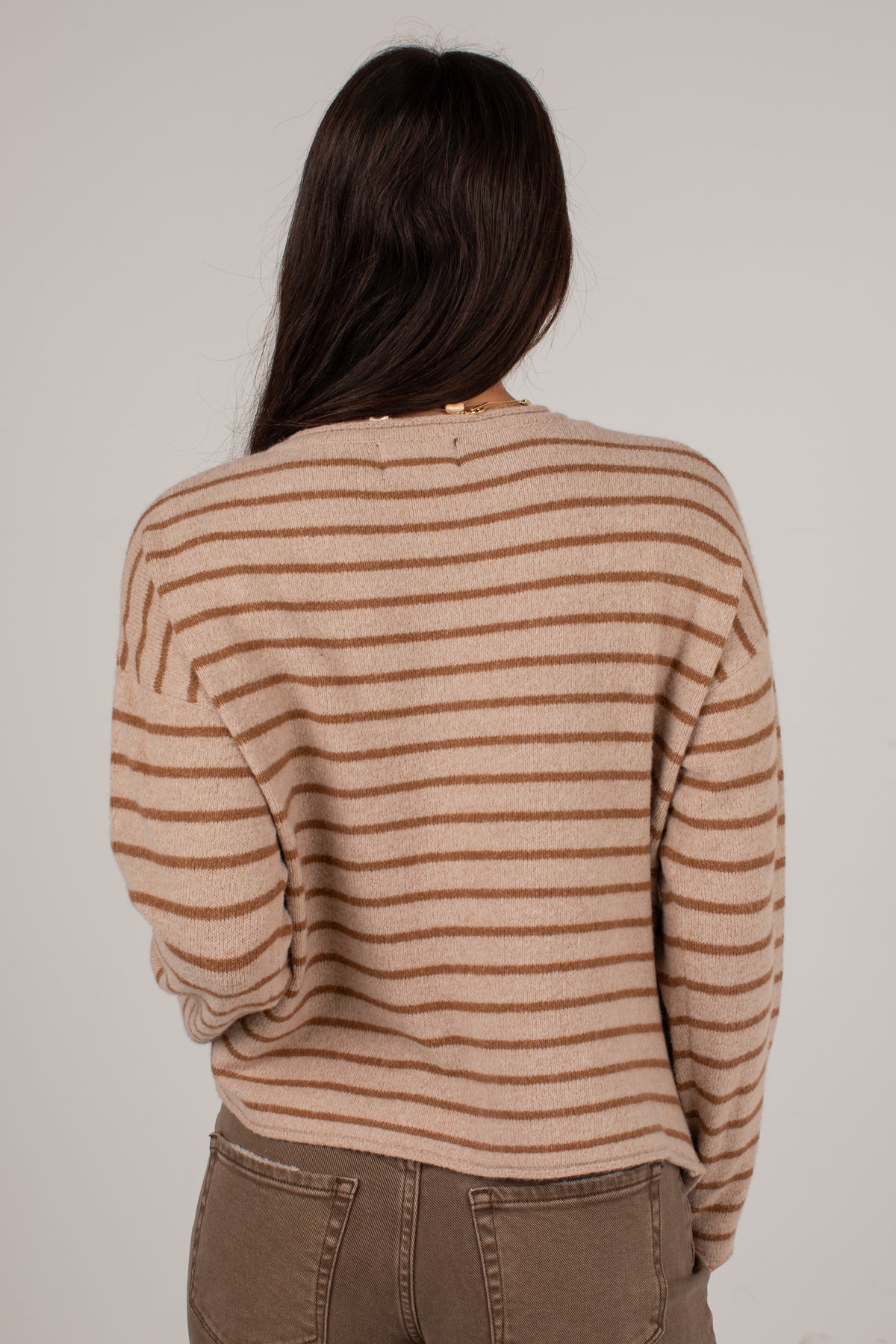 A woman viewed from behind wearing a beige and brown horizontally striped knit sweater and brown pants against a plain light gray background.