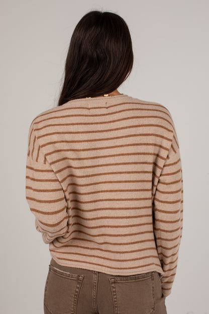 A woman viewed from behind wearing a beige and brown horizontally striped knit sweater and brown pants against a plain light gray background.