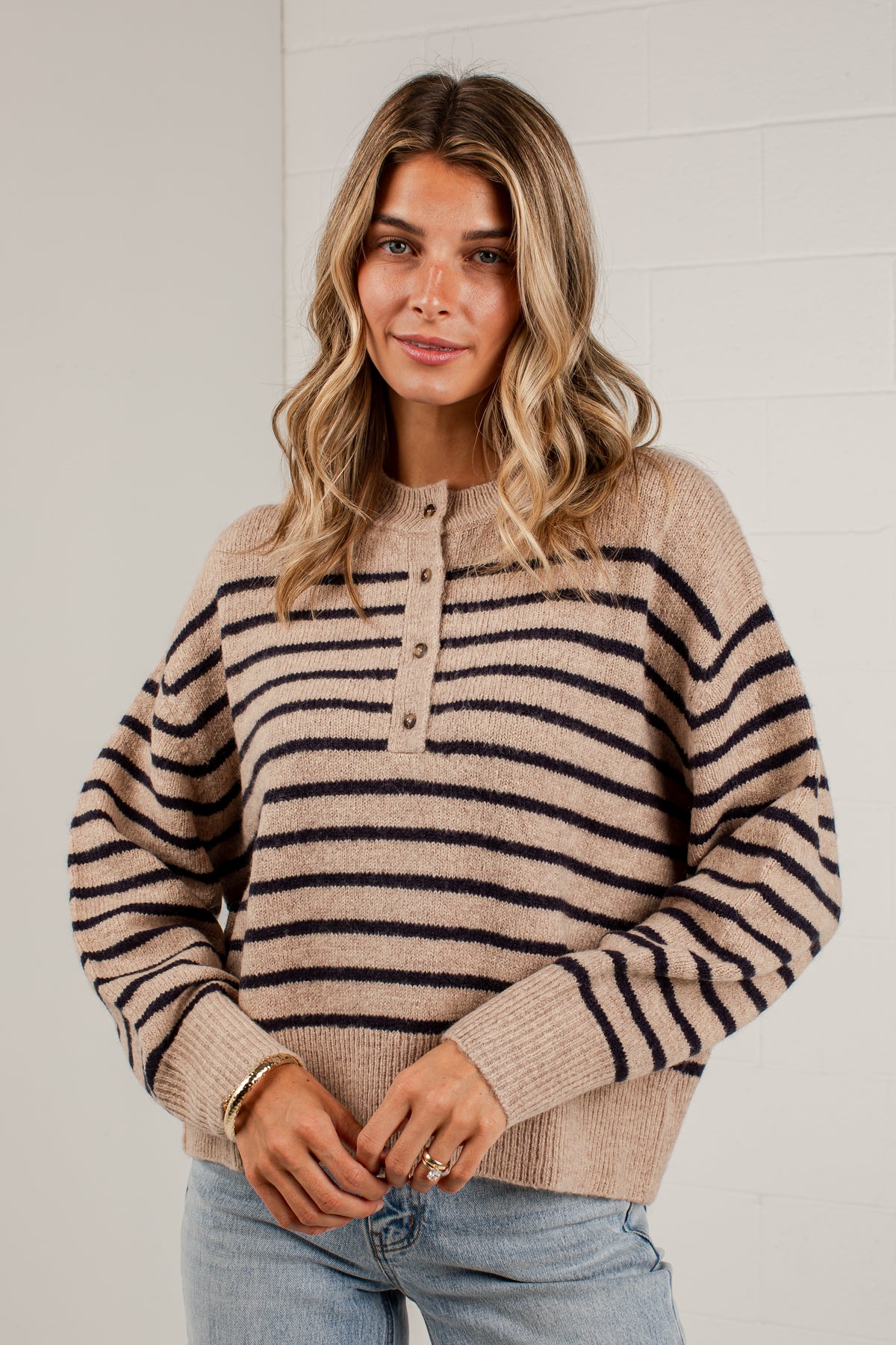 A woman wearing a beige and black horizontally striped knit henley sweater with a buttoned collar, paired with light blue jeans, standing against a white brick wall.