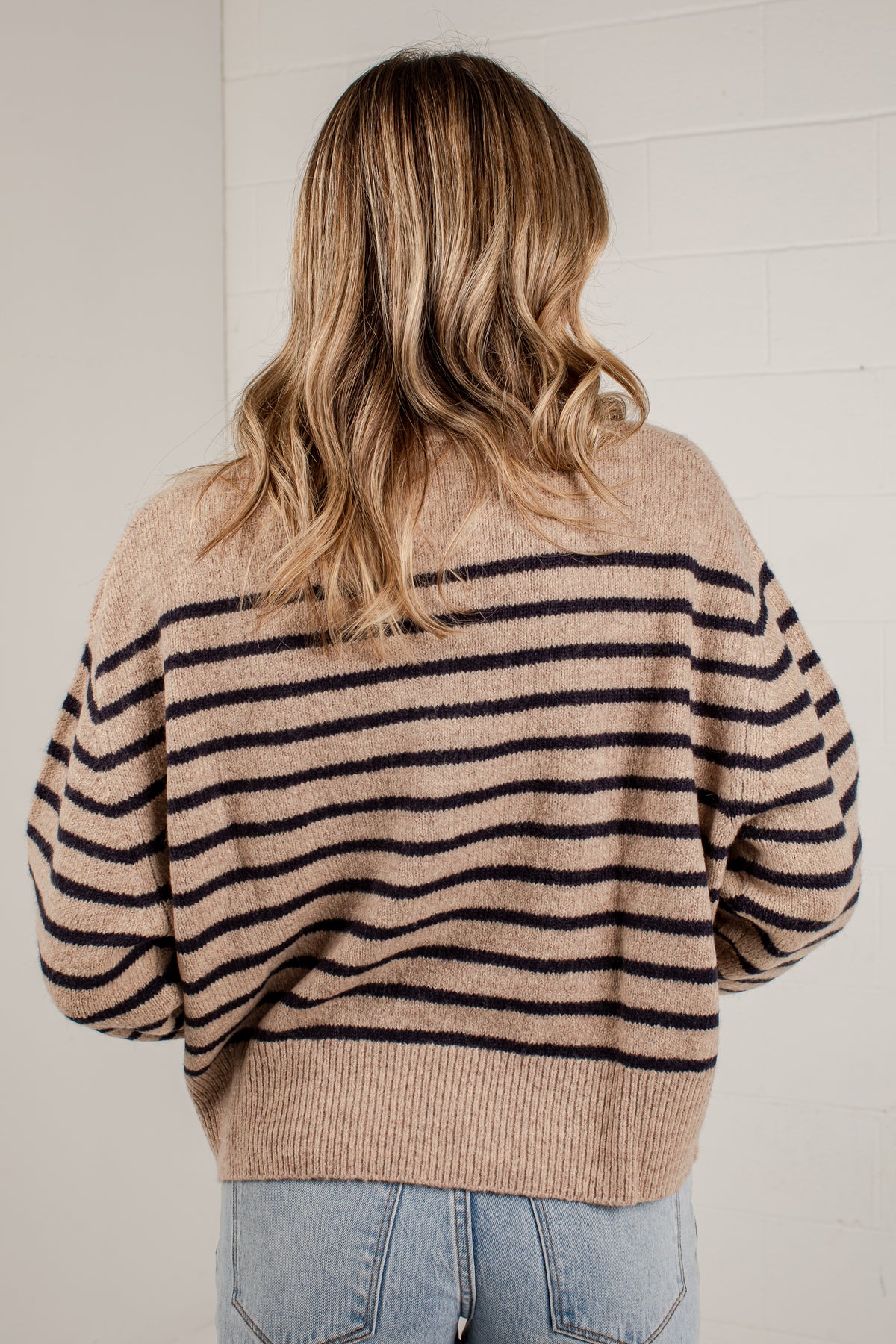 A woman viewed from behind wearing a beige knit sweater with horizontal black stripes and light blue jeans, standing against a white brick wall.
