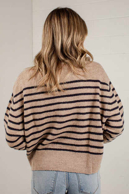 A woman viewed from behind wearing a beige knit sweater with horizontal black stripes and light blue jeans, standing against a white brick wall.