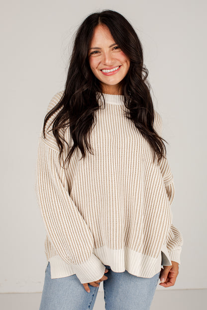 A smiling woman with long dark hair wearing an oversized cream and beige vertically striped knit sweater and light blue jeans, standing against a plain light gray studio background.