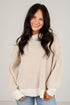 A smiling woman with long dark hair wearing a cream and beige vertically ribbed knit sweater with balloon sleeves, paired with light blue jeans, against a plain light gray background.