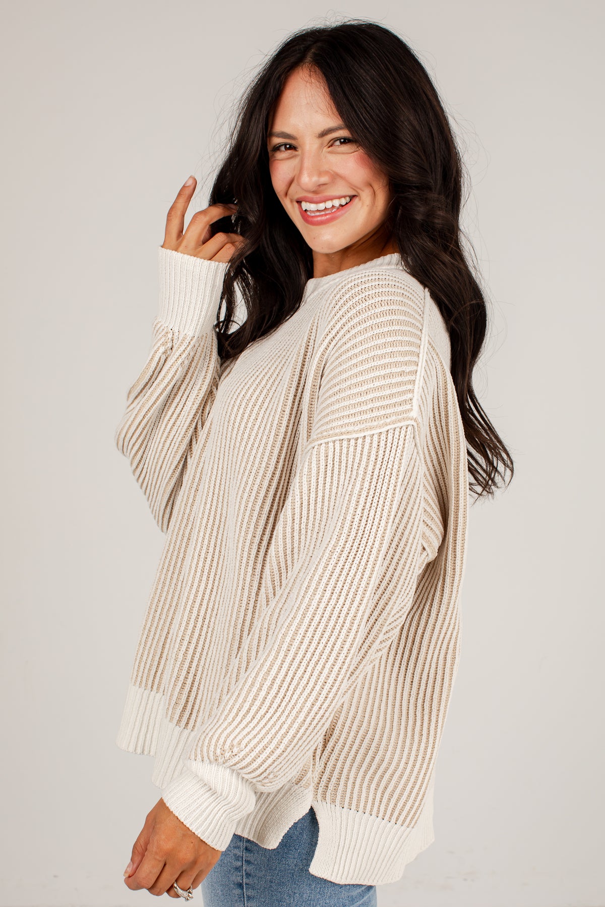 A woman wearing a cream and beige vertically striped knit sweater with ribbed cuffs and hem, paired with blue jeans, photographed against a light gray studio background.