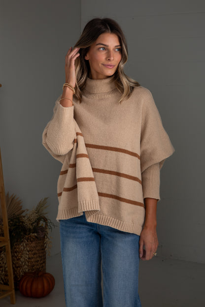 Woman wearing a beige poncho with brown stripes indoors against a gray wall.
