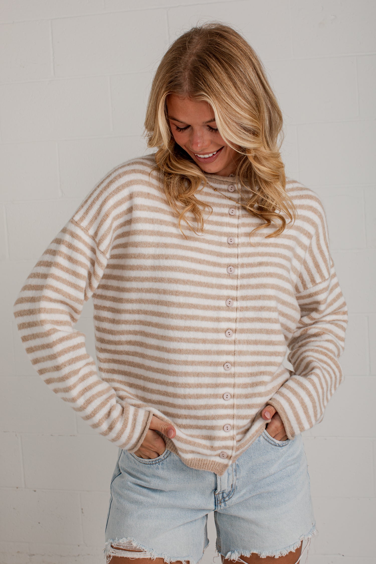 A woman wearing a beige and white striped button-up knit sweater and light-wash denim shorts, standing against a white brick wall.