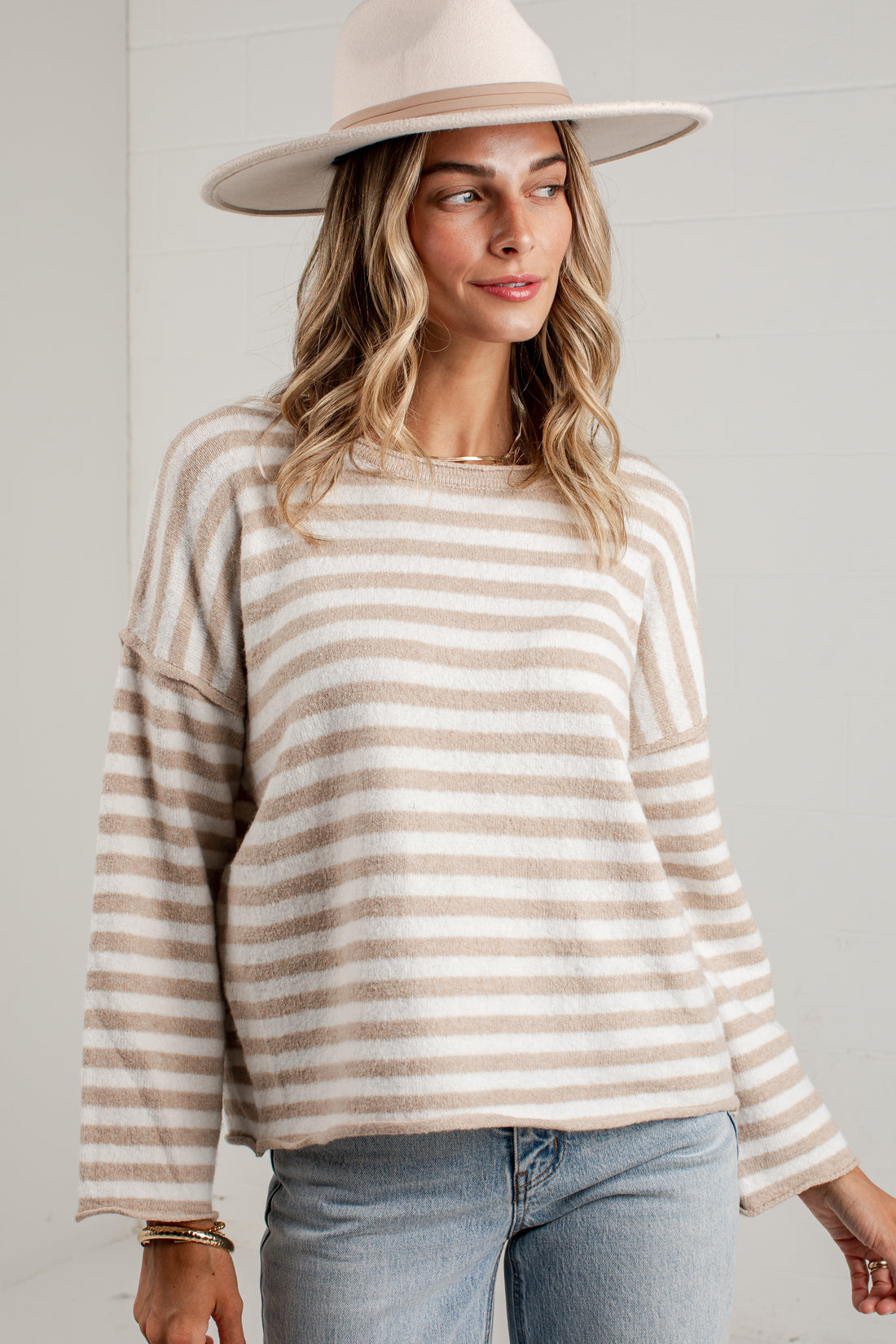A woman wearing a beige and white striped long-sleeve knit sweater, a cream wide-brim hat, and light blue jeans, standing against a white paneled wall.