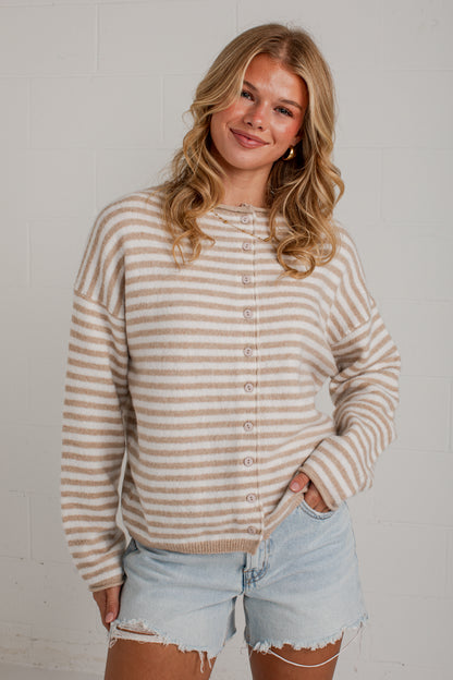 A woman wearing a beige and white striped button-up cardigan sweater and light-wash denim shorts, standing against a white brick wall.