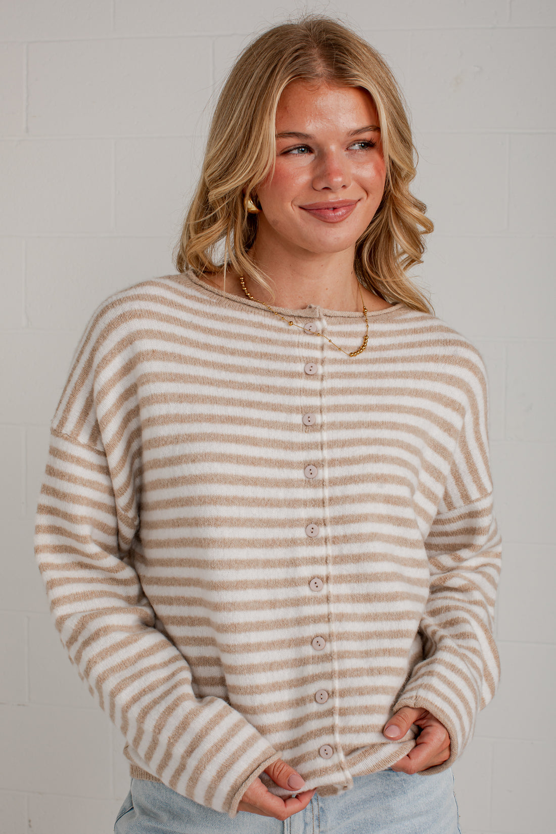A woman wearing a beige and white horizontally striped button-up cardigan sweater, paired with light blue jeans, standing against a white brick wall.