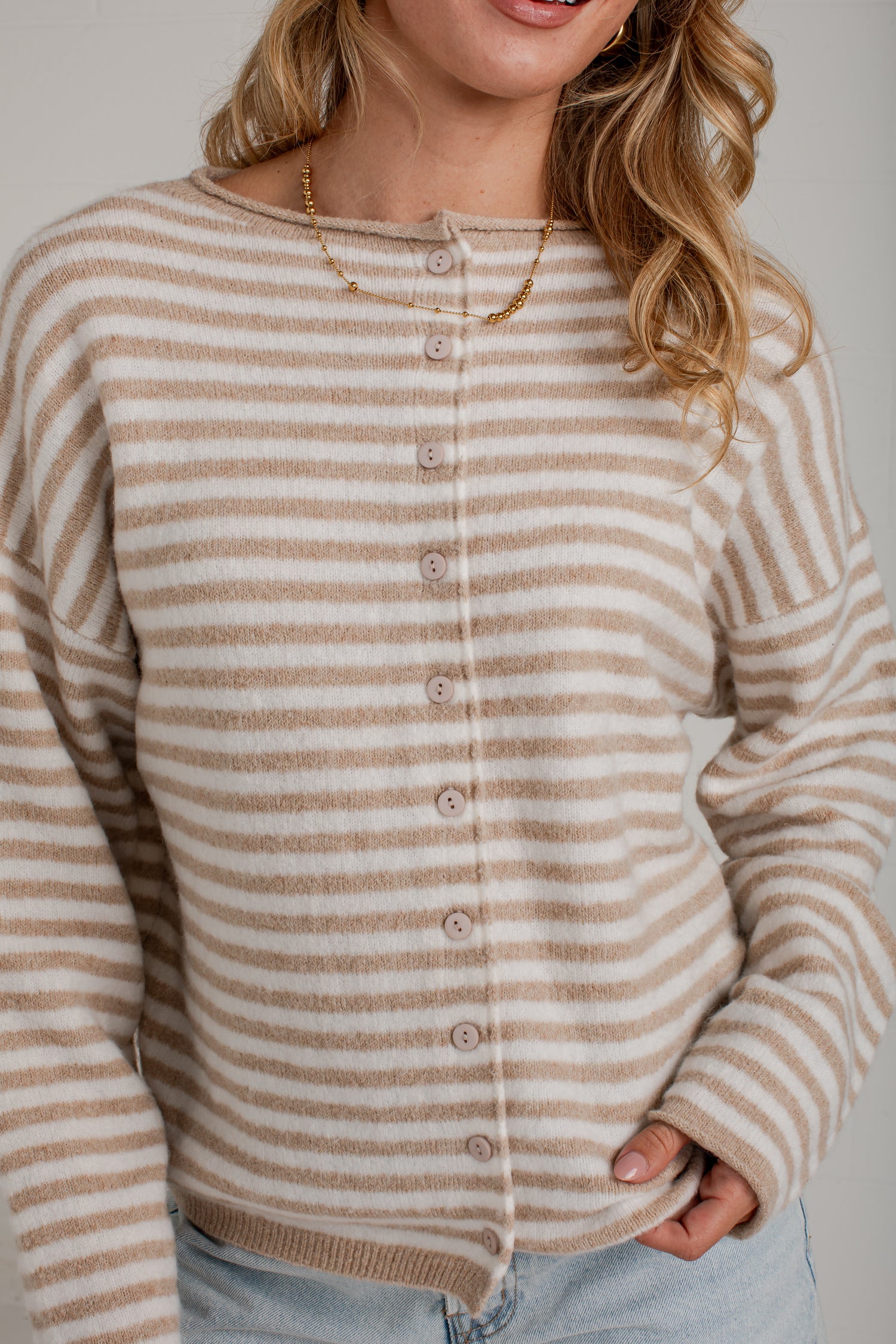 A woman wearing a beige and white horizontally striped button-up cardigan sweater, paired with light blue jeans and a gold necklace, against a neutral gray background.