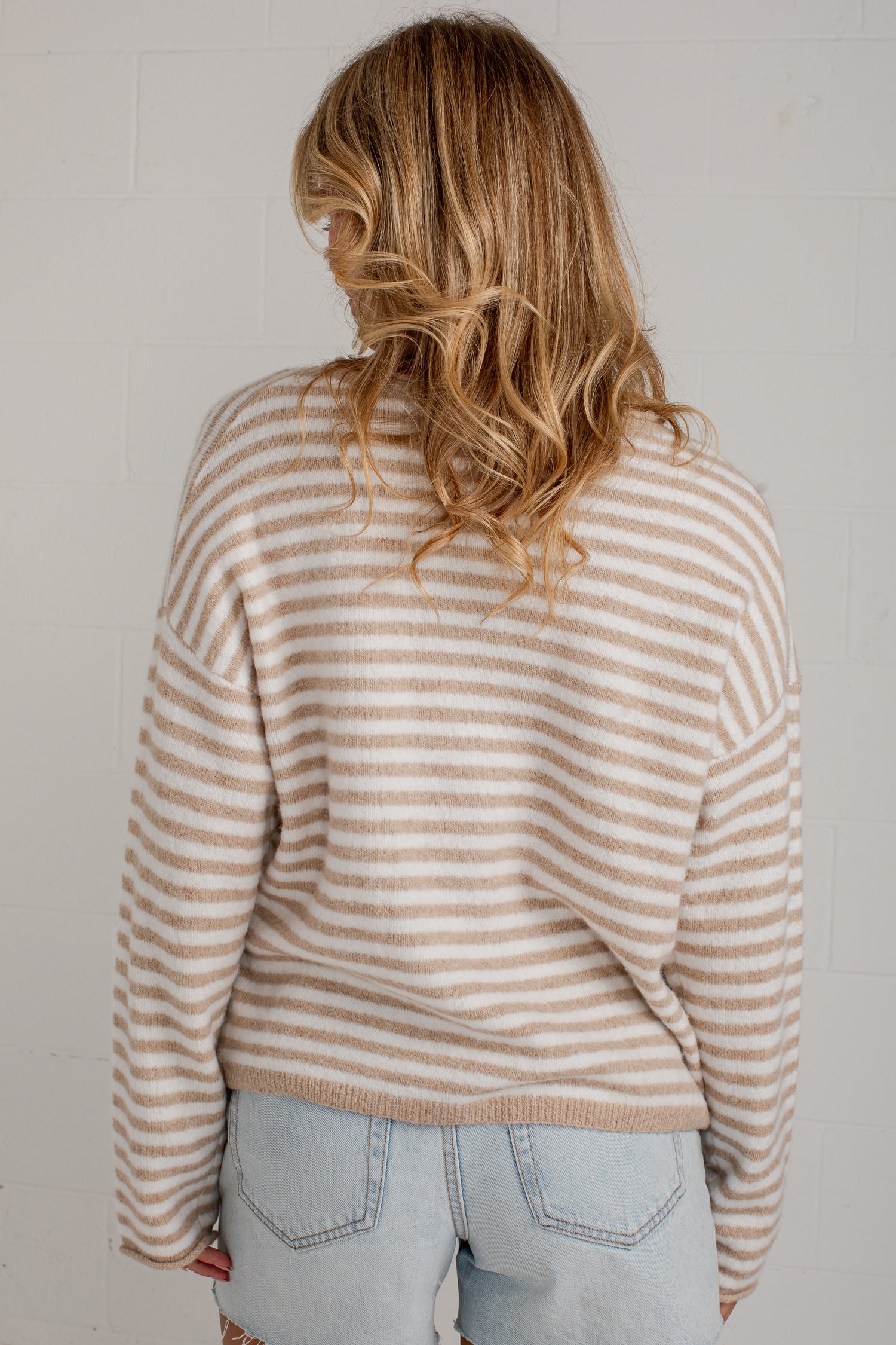 A woman viewed from behind wearing a beige and white horizontally striped knit sweater and light blue jeans against a white brick wall.