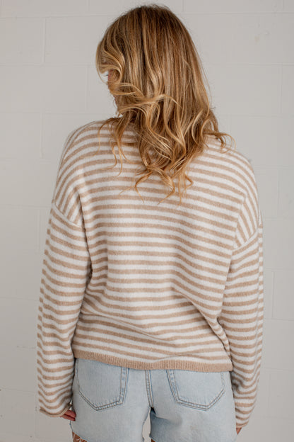 A woman viewed from behind wearing a beige and white horizontally striped knit sweater and light blue jeans against a white brick wall.