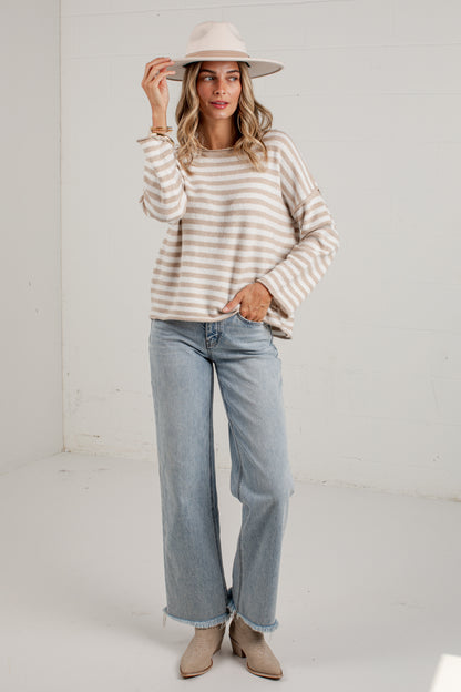 A woman modeling a beige and white striped long-sleeve knit sweater, paired with light-wash wide-leg jeans and beige ankle boots, standing against a white studio wall.