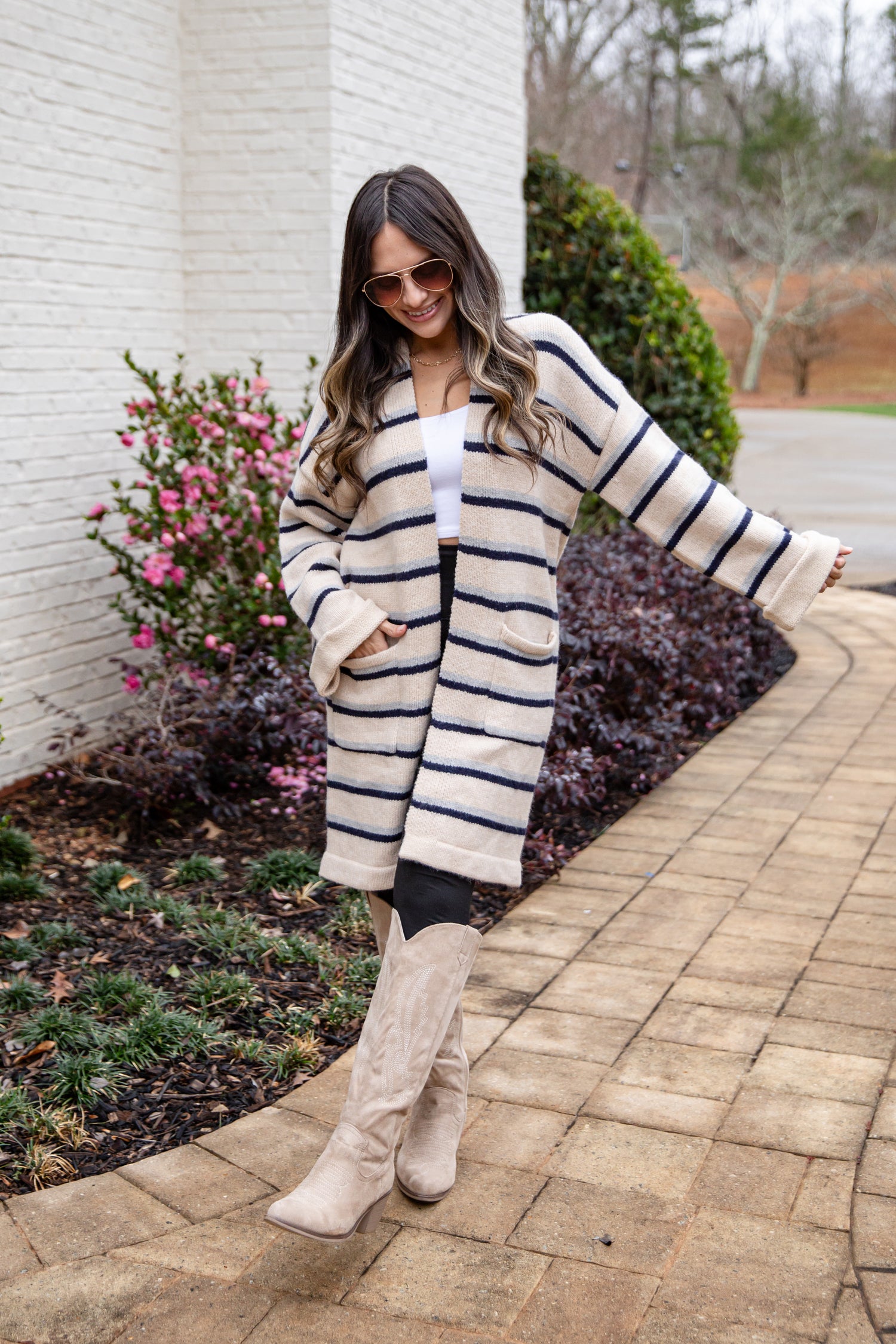 Woman wearing a striped cardigan and white boots standing on a sidewalk with plants and flowers in the background.