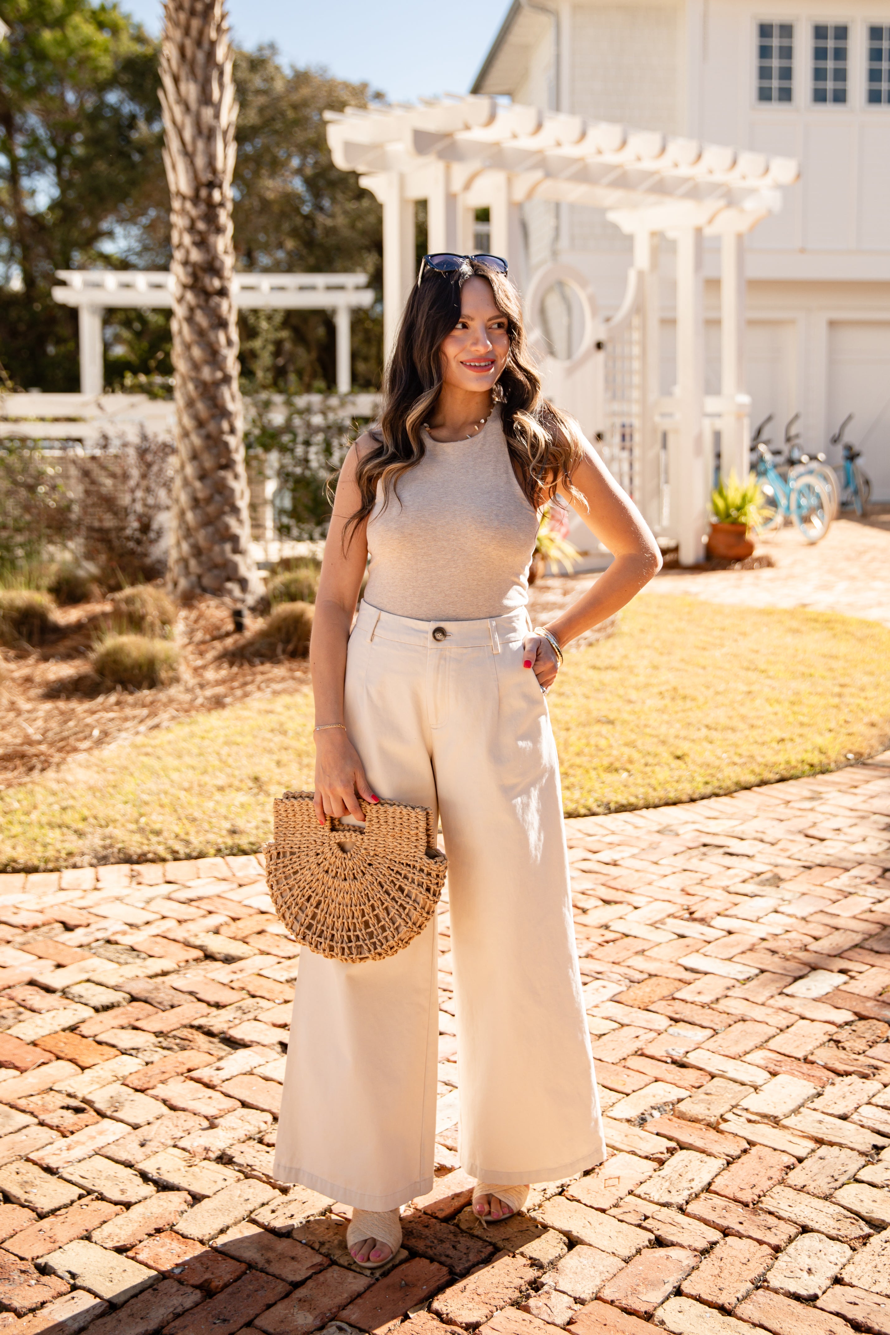 Woman in a beige top and white pants standing on a brick path with a white building and trees in the background.