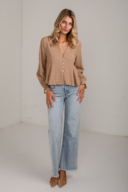 A woman modeling a light brown long-sleeve button-up blouse with a peplum hem and light-wash wide-leg jeans, standing against a plain white studio wall.
