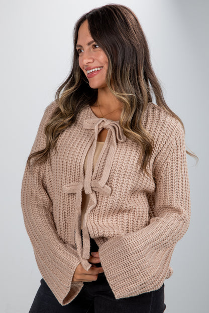 Woman wearing a beige knitted sweater with a tie detail on a plain background