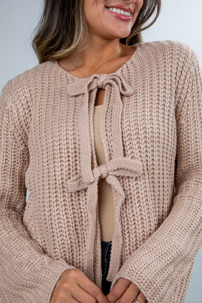 Woman wearing a beige knitted cardigan with a bow detail on a plain background